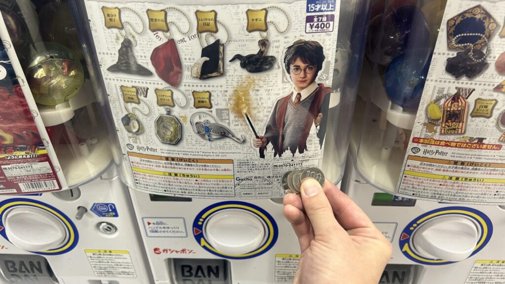 Hand holding four 100-yen coins ready to play a Harry Potter themed Gachapon machine priced at 400 yen